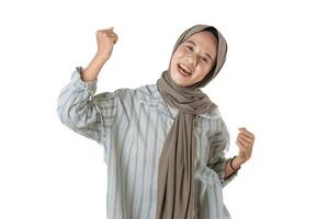 A cheerful woman wearing a grey hijab and a striped shirt raises her fists in excitement, expressing joy and positive energy against a white background. photo