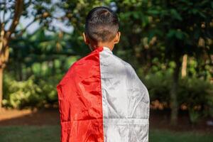 A child is seen from behind, wrapped in an Indonesian flag, standing outdoors with a background of green trees and plants, symbolizing national pride. photo