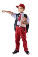 A young Indonesian boy in a red and white school uniform points forward while holding a book and wearing a backpack. photo