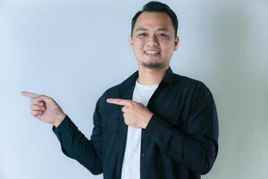 A man in a black shirt, white t-shirt, and short hair is smiling and pointing with both hands to the left, against a plain white background. photo
