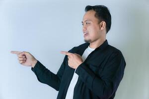 A man in a black shirt, white t-shirt, and short hair is smiling and pointing with both hands to the left, against a plain white background. photo