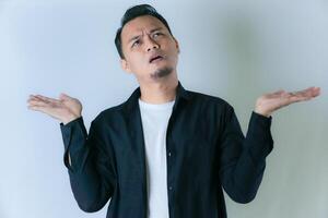 A man in a black shirt and white t-shirt looks confused, raising both hands with a questioning expression against a plain white background. photo