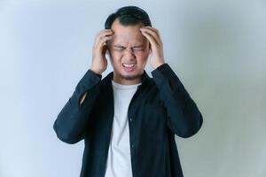 A man in a black shirt and white undershirt is holding his head, appearing to be in pain or distress, against a plain light background. photo