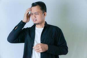A man in a black shirt and white undershirt is holding his head, appearing to be in pain or distress, against a plain light background. photo