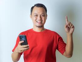 A man in a red shirt smiles while holding a smartphone in his right hand and pointing upwards with his left index finger against a plain background. photo
