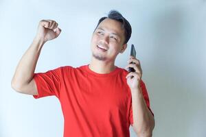 A man in a red shirt is seen celebrating with a joyful expression, raising his fist while holding a phone, perhaps indicating good news or a victory. photo