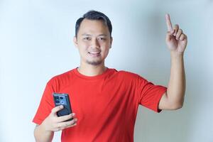 A man in a red shirt smiles while holding a smartphone in his right hand and pointing upwards with his left index finger against a plain background. photo