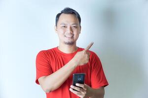 A man in a red shirt smiles while holding a smartphone in his left hand and pointing upwards with his right index finger against a plain background. photo
