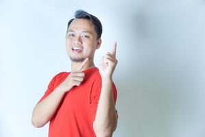 A cheerful man in a red shirt poses against a white background, smiling confidently while pointing to his side with both hands. photo