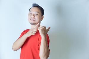 A cheerful man in a red shirt poses against a white background, smiling confidently while pointing to his side with both hands. photo