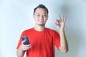 A man in a red shirt holds a smartphone in one hand, giving an OK hand gesture with a smile, indicating satisfaction or approval. photo