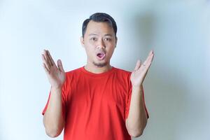 A man in a red shirt stands against a plain background, expressing surprise with wide eyes and open mouth, his hands raised on either side of his head. photo