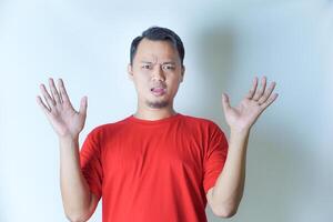 A man in a red shirt stands against a plain background, expressing surprise with wide eyes and open mouth, his hands raised on either side of his head. photo
