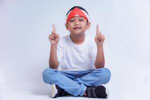 A boy wearing a white shirt and jeans sits cross-legged, pointing up with both hands, smiling brightly. photo