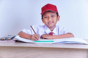 A Indonesian young boy in a school uniform is sitting at a desk, smiling while drawing on a piece of paper with colored pencils beside him. photo