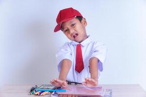 An Indonesian boy in school uniform points to a book on a table with an expression of refusal to learn photo
