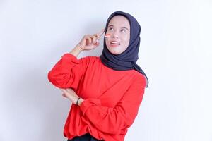 A young Indonesian woman wearing a hijab, pointing her right index finger towards her temple, appears to be thinking or as if having an idea photo