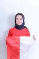 A young Indonesian woman wearing a hijab holds a red and white flag, with matching colored stripes on her cheeks. She smiles and looks directly at the camera, isolated against a white background. photo