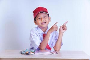 An Indonesian boy in a school uniform smiles and points upwards, sitting in front of a desk with books and crayons, wearing a red hat. photo