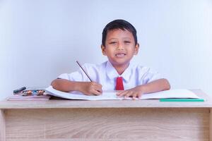 A Indonesian young boy in a school uniform is studying at a desk with pencils and books. photo