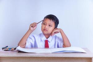 A Indonesian young boy in a school uniform looks thoughtful with a pencil to his head, sitting at a desk with an open notebook and colors. photo