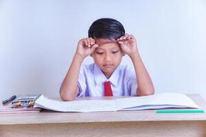 A Indonesian young boy in a school uniform looks contemplative with a pencil on his forehead, sitting at a desk with books and crayons. photo