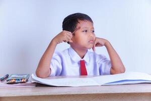 A Indonesian young boy in a school uniform looks thoughtful with a pencil to his head, sitting at a desk with an open notebook and colors. photo