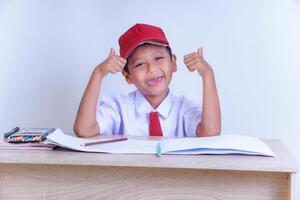 An Indonesian boy in school uniform sits in front of a wooden desk wearing a white shirt red tie and red hat. He looks cheerful, making funny gestures with his two thumbs. Isolated on white background photo