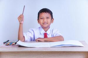 A Indonesian young boy in a school uniform, sitting at a desk with a raised hand, smiling, with books and crayons, ready to learn. photo