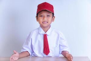 An Indonesian boy in a school uniform is smiling, sitting at a desk with both hands resting on it. He is wearing a white shirt, a red tie, and a red cap. A plain, bright background highlights him. photo