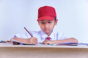 An Indonesian boy in school uniform is focused on writing in a notebook on a table, with a red hat. photo