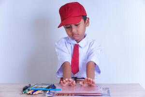 A Indonesian young boy in a school uniform sits at a desk, with a red cap and tie, looking at books and colorful pencils on the table. photo
