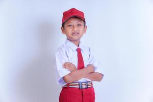 A Indonesian young boy in a school uniform, with a red cap and tie, stands smiling confidently with arms crossed on a white background. photo