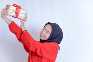 A young woman in a hijab smiled while holding a gift wrapped in shiny gold paper with a red ribbon, looking at it joyfully. celebrating Indonesia's independence day on August 17. photo