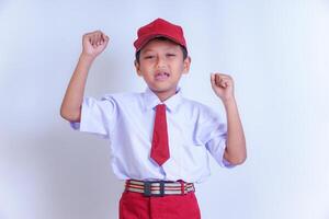 A Indonesian young boy in a school uniform poses confidently with both fists raised in the air. He is wearing a white shirt, red tie, and red cap. Isolated on white background photo