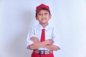 A Indonesian young boy in a school uniform, with a red cap and tie, stands smiling confidently with arms crossed on a white background. photo