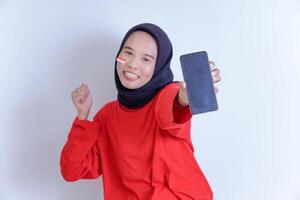 A young Indonesian woman wearing a hijab is smiling brightly while holding a phone and showing the national flag on her cheek, wearing a vibrant red shirt, isolated on white background photo