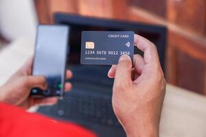 A person holding a credit card in one hand and a smartphone in the other while near a laptop. photo