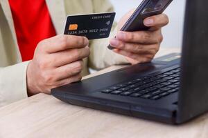 A person with red shirt holding a credit card in one hand and a smartphone in the other while near a laptop. photo