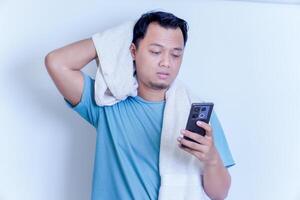 an Asian man with a disappointed expression while holding his smartphone and a towel on his shoulder photo