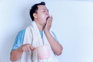 A Asian man yawns while holding a pink dipper, wrapped in a towel, looking sleepy. photo