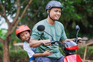 A asian father and son smiling together on a electric scooter, both wearing helmets in a green setting. photo