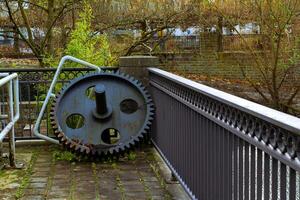 A large, rusty gear leans against a railing in a quiet, overgrown photo