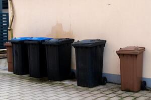 Various trash bins in different colors are lined up against a building photo