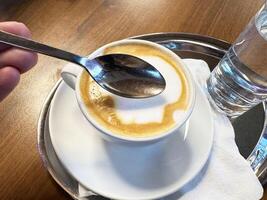 Hand holding spoon above cappuccino on wooden table with glass of water photo