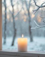 Cozy winter scene with lit candle and snow covered trees outside a window photo