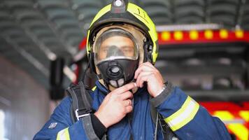 Male firefighter taking off helmet against background of a fire engine. Portrait of young fireguard in protective uniform after fire near big red car at department. Concept of heroic profession video