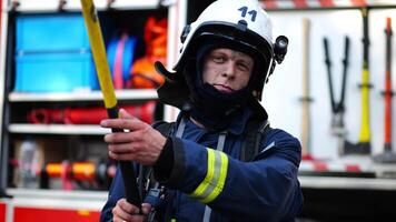 Portrait of confident male fireguard with axe standing near a fire engine. Young fireman with dirty face looking at camera in full equipment after fire against the background of big red truck video