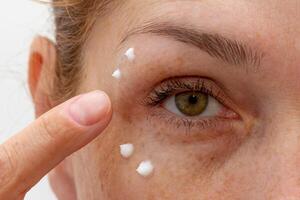 Close up cropped eye of middle aged caucasian woman looking at camera applying cream on skin photo
