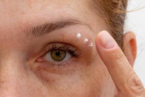 Close up cropped eye of middle aged caucasian woman looking at camera applying cream on skin photo